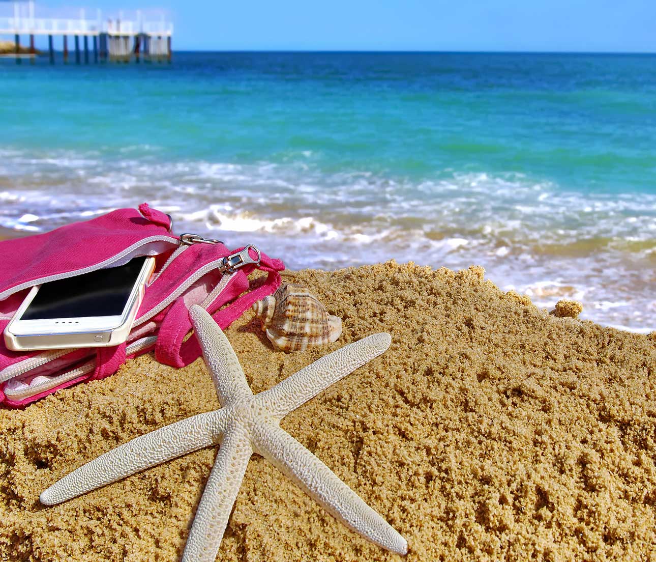 A smart phone in a pink beach bag on the beach next to a starfish and the ocean.